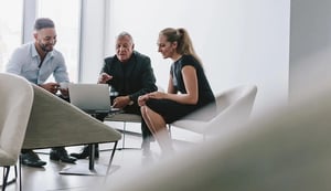 woman sitting in white chair leaning over to look at laptop with two men pointing to screen